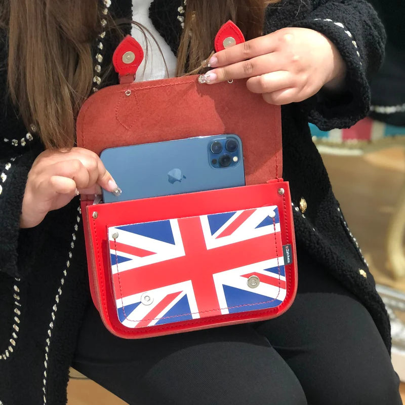 A woman putting an iPhone in a midi satchel bag, in red, featuring the Union Jack on the front pocket